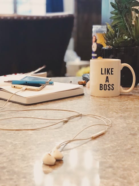 A close-up view of a cluttered office desk surface featuring a white ceramic mug with black text that reads 'LIKE A BOSS', placed towards the right side of the image. To the left of the mug, there are several closed notebooks or planners stacked, with a blue pen resting on top, and a pair of white earphones with a tangled cord lying on the desk. In the background, part of a black office chair is visible, along with a blurred shelf holding various items, including a small potted plant with green foliage and a jar or container. The desk appears to be made of a light-colored, smooth surface, possibly wood or laminate. The natural ambient lighting highlights the texture of the desk and objects, creating a professional yet informal workspace scene that can relate to office environments where waste or clutter management might be considered, and the mug's message adds a casual, motivational touch.