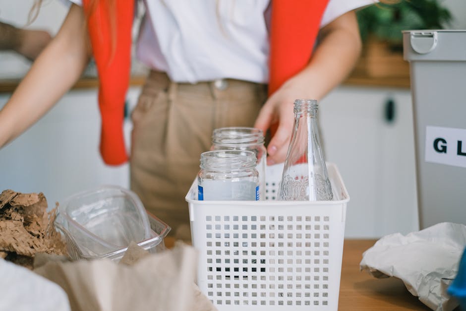 A person wearing a white shirt and beige trousers is placing three empty glass jars into a white plastic basket with a grid design, situated on a wooden table. The environment appears to be an indoor recycling or waste collection area, with additional transparent containers and brown paper packaging visible in the foreground, and a grey waste bin or storage unit partially visible to the right, with a label that suggests waste separation. The background includes slightly blurred elements of indoor furniture and natural lighting, creating a clean and organized space dedicated to recycling or rubbish sorting activities. The scene reflects an aspect of private waste handling and eco-friendly disposal, consistent with rubbish collection services and environmentally conscious waste management practices, as operated by companies such as Rubbish Clearance Lewisham.