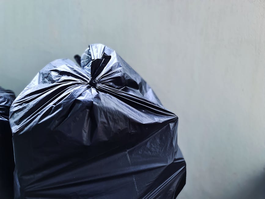 Close-up of a large black rubbish bag made of shiny, plastic material with visible creases and folds, positioned in the foreground on the left side of the image. The bag is tightly tied at the top, with the gathered plastic creating a puckered effect, and appears to contain waste, possibly household rubbish. In the background, a portion of another similar black bag is partially visible, slightly out of focus. The setting features a plain, light-colored wall with a smooth texture, and the lighting is neutral, illuminating the bags clearly without harsh shadows. The scene suggests an indoor environment, such as a storage area or utility space, where waste is temporarily stored before disposal, aligning with the context of private rubbish collection and on-site clearance services offered by Rubbish Clearance Lewisham.