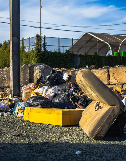 A pile of mixed waste rubbish is scattered on a gravel surface in an outdoor setting, consisting of black plastic garbage bags, a yellow plastic container, a worn beige car seat with fabric upholstery, and various loose debris. Behind the rubbish, a low stone wall separates the area from a green hedge and some tall trees, with an industrial or sports facility with a curved, semi-transparent roof visible in the background. A utility pole with cables extends vertically on the left side of the image, under a blue sky with some wispy clouds. The scene appears to depict a temporarily abandoned rubbish accumulation, possibly part of an independent waste removal effort, with natural light illuminating the scene, supporting the context of private rubbish clearance services by companies like Rubbish Clearance Lewisham.