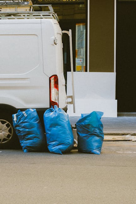 Close-up of a large black rubbish bag made of shiny, plastic material with visible creases and folds, positioned in the foreground on the left side of the image. The bag is tightly tied at the top, with the gathered plastic creating a puckered effect, and appears to contain waste, possibly household rubbish. In the background, a portion of another similar black bag is partially visible, slightly out of focus. The setting features a plain, light-colored wall with a smooth texture, and the lighting is neutral, illuminating the bags clearly without harsh shadows. The scene suggests an indoor environment, such as a storage area or utility space, where waste is temporarily stored before disposal, aligning with the context of private rubbish collection and on-site clearance services offered by Rubbish Clearance Lewisham.