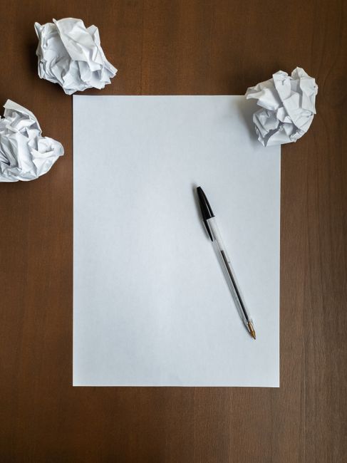 A blank white sheet of paper lies flat on a wooden surface, with a black ballpoint pen resting diagonally across the center of the sheet. Surrounding the paper are three crumpled balls of white paper, placed at the top left, top right, and bottom left corners of the image. The scene appears to be set in an indoor environment with even, natural lighting, creating clear shadows around the crumpled paper. The overall composition suggests a workspace or desk area where ideas or plans might be in development, relevant to discussions about avoiding common mistakes in private rubbish disposal or independent waste clearance, as provided by companies like Rubbish Clearance Lewisham.