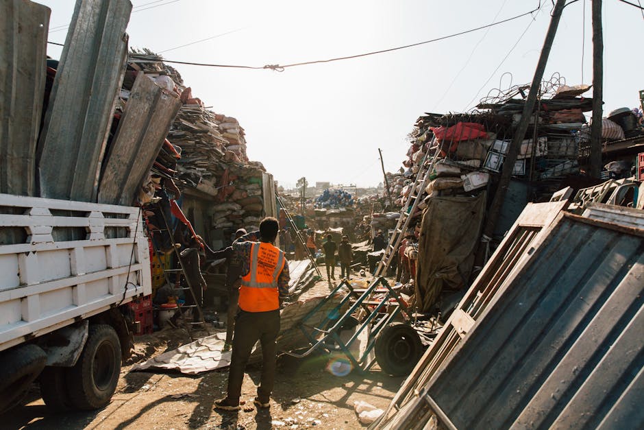 A scene at a densely cluttered outdoor rubbish dump, with tall stacks of mixed waste materials including wooden planks, metal sheets, cardboard boxes, and plastic containers on either side of a narrow pathway. The surfaces of the waste are weathered and vary in texture from rough wood to smooth metal, with colors predominantly in shades of brown, grey, and black, with splashes of red and other muted tones. Several individuals are present; one person wearing an orange high-visibility vest is standing in the foreground, overseeing the scene, while others are seen in the background working amidst the pile, possibly involved in rubbish collection or clearance activities. A white truck with an open cargo area is parked on the left side, partially visible, suggesting the collection or transportation of waste. The environment appears sunlit with a clear sky, and overhead wires cross the scene, indicating an industrial or urban setting. This image illustrates the kinds of refuse and materials managed by private waste removal services like Rubbish Clearance Lewisham, often involved in on-site clearance and alternative waste handling solutions.