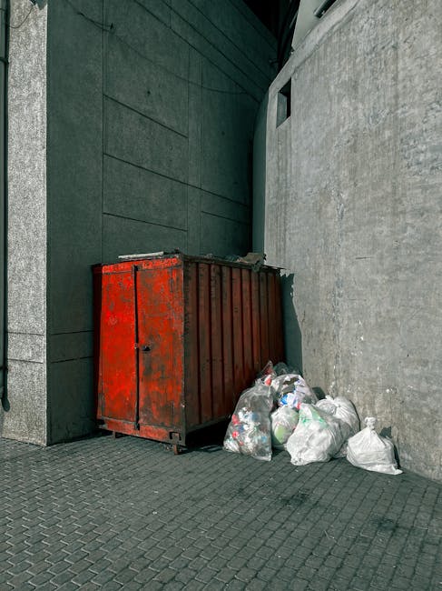 Three large black plastic rubbish bags, filled and tied, rest upright on a grey concrete sidewalk near a black metal fence with vertical bars, set against a background of dense green foliage and a dark wooden structure. The bags appear to contain household waste materials, possibly including packaging or soft refuse, with some bulging and showing some slight creases on their surfaces. The scene is outdoors, illuminated by natural ambient light, casting minimal shadows and highlighting the textured surfaces of the plastic bags. The overall setting suggests a typical private property or urban street environment where waste has been temporarily collected, possibly awaiting collection or disposal by a rubbish clearance service such as Rubbish Clearance Lewisham, highlighting the everyday process of private waste handling outside of official bin collections. The surrounding elements and arrangement emphasize a straightforward scene of waste awaiting removal, consistent with non-regular or alternative rubbish clearance options.