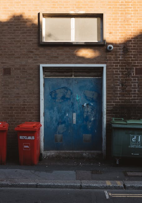 The image depicts a brick building exterior with a large, weathered blue metal door positioned centrally on the wall. Above the door, there is a rectangular window with frosted or dirty glass panes framed by black metal, allowing some diffused light to enter. To the right of the window, a small black security camera or sensor is mounted on the brick wall. In front of the building, on the pavement, there are three visible waste containers: two red recycling bins made of plastic with lids, and one green bin, likely for general waste, also made of plastic. The red bins are labeled 'RECYCLABLES,' while the green bin has some text and a logo. The scene is illuminated by natural daylight, casting a shadow of a tree or nearby object onto the brick wall. The overall context suggests an urban setting where waste management and disposal are handled through external bins placed outside the building, reflecting typical arrangements for independent rubbish collection outside commercial or industrial premises, possibly related to private waste handling services such as those provided by Rubbish Clearance Lewisham.