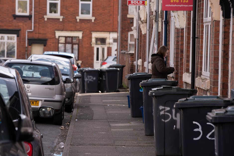 A woman standing on a sidewalk next to a row of black recycling bins and waste containers positioned against the brick exterior of a residential building. The bins are made of plastic with lids, some of which are closed, and are marked with white chalk or spray paint. The woman, wearing a dark brown jacket with a hood, appears to be handling one of the bins. In the background, several parked cars line the street, including a small silver hatchback closest to the camera. The street is lined with red brick terraced houses that have white-framed windows and small front gardens or steps leading to entrances. The scene is lit with natural daylight, and a red sale sign is partially visible above the woman, attached to the building facade. The environment suggests an urban area where private waste collection and rubbish clearance might occur, supporting services offered by companies like Rubbish Clearance Lewisham.