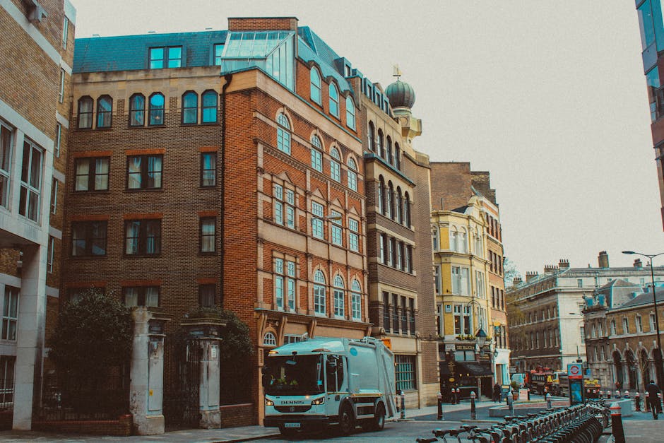 The image shows a street scene in an urban area with a mix of historic and modern buildings. In the foreground, a light blue refuse collection vehicle is parked along the curb, partially blocking the view of a historic brick building with multiple stories. The brick building features large, arched windows with blue frames, and a decorative cornice along the roofline. Adjacent buildings include a yellow Victorian-style structure with detailed window frames and a rounded turret, as well as contemporary glass and steel structures on either side. The street is lined with black bollards and bicycles parked along the pavement, with a few pedestrians visible in the background. Overhead, a cloudy sky casts diffuse lighting, creating a neutral atmosphere. The scene subtly relates to independent waste management services, highlighting the vehicle as part of the private rubbish collection processes that complement alternative waste handling solutions in urban environments, with Rubbish Clearance Lewisham nearby for rubbish disposal needs.
