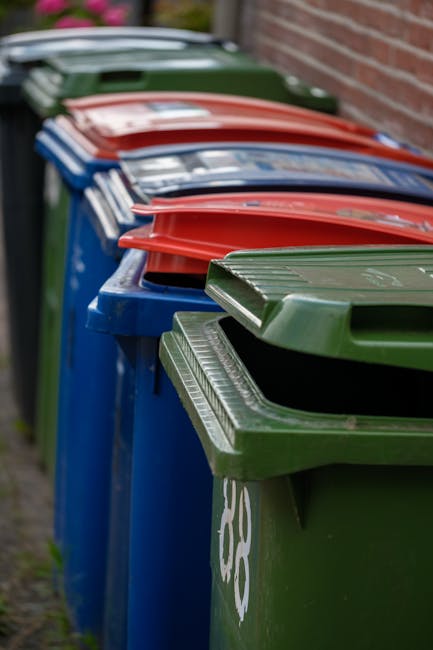 A row of multiple outdoor wheelie bins positioned along a pavement beside a brick wall, with the bins arranged in a linear formation. The bins are in different colours, including blue, green, red, and dark green, each with a textured plastic lid featuring ridged patterns. The lids are closed, and the bins appear to be for household waste disposal, typically associated with private waste collection services such as those offered by Rubbish Clearance Lewisham. The bins are situated outdoors, likely in a residential area, on a concrete or paved surface, and are lit by natural daylight, creating subtle shadows on the ground. The background shows a blurred view of a garden or driveway area, emphasizing the focus on the bins' alignment and condition, which is typical in rubbish removal or collection scenarios, supporting the context of alternative waste handling methods outside of local authority services.