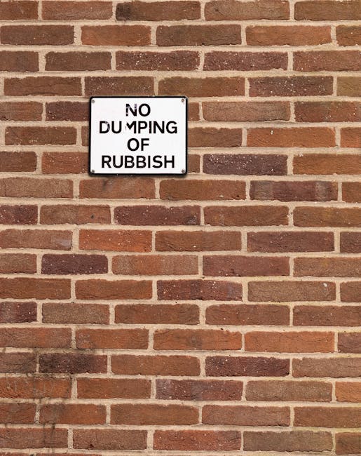 A close-up view of a rectangular metal sign mounted on a red brick wall. The sign displays the message 'NO DUMPING OF RUBBISH' in black uppercase letters on a white background, with some of the letters slightly faded or broken. The brick wall features evenly spaced, horizontally arranged bricks with a reddish-brown and dark brown color palette, and light-colored mortar joints. The shadows cast on the wall suggest natural outdoor lighting. The sign indicates restrictions related to waste disposal, supporting the context of rubbish removal and private waste management in the area.