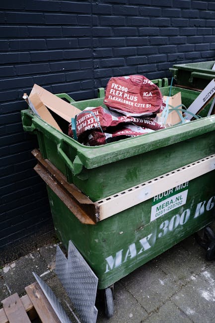 A row of multiple outdoor wheelie bins positioned along a pavement beside a brick wall, with the bins arranged in a linear formation. The bins are in different colours, including blue, green, red, and dark green, each with a textured plastic lid featuring ridged patterns. The lids are closed, and the bins appear to be for household waste disposal, typically associated with private waste collection services such as those offered by Rubbish Clearance Lewisham. The bins are situated outdoors, likely in a residential area, on a concrete or paved surface, and are lit by natural daylight, creating subtle shadows on the ground. The background shows a blurred view of a garden or driveway area, emphasizing the focus on the bins' alignment and condition, which is typical in rubbish removal or collection scenarios, supporting the context of alternative waste handling methods outside of local authority services.