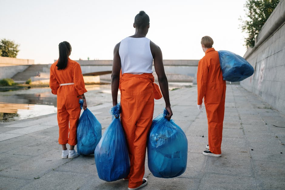 Three waste collection workers dressed in bright orange uniforms are walking along a paved pathway beside a body of water, each carrying large blue refuse bags filled with rubbish. The worker in the foreground, a woman with dark skin and her hair tied up in a bun, holds two bags, one in each hand, with a textured, slightly crinkled plastic surface. To her left, another woman with long dark hair tied back is also carrying a bag, while on the right, a man with light hair and fair skin carries a large blue bag slung over his shoulder. Behind them, a concrete embankment or retaining wall runs parallel to the pathway, with some greenery visible at the top. The scene is illuminated by natural daylight, creating a neutral, professional atmosphere typical of outdoor waste collection activities, reflecting an alternative approach to rubbish removal that involves on-site or private collection services. The image visually supports the context of rubbish clearance and waste management, emphasizing proper disposal and collection practices.