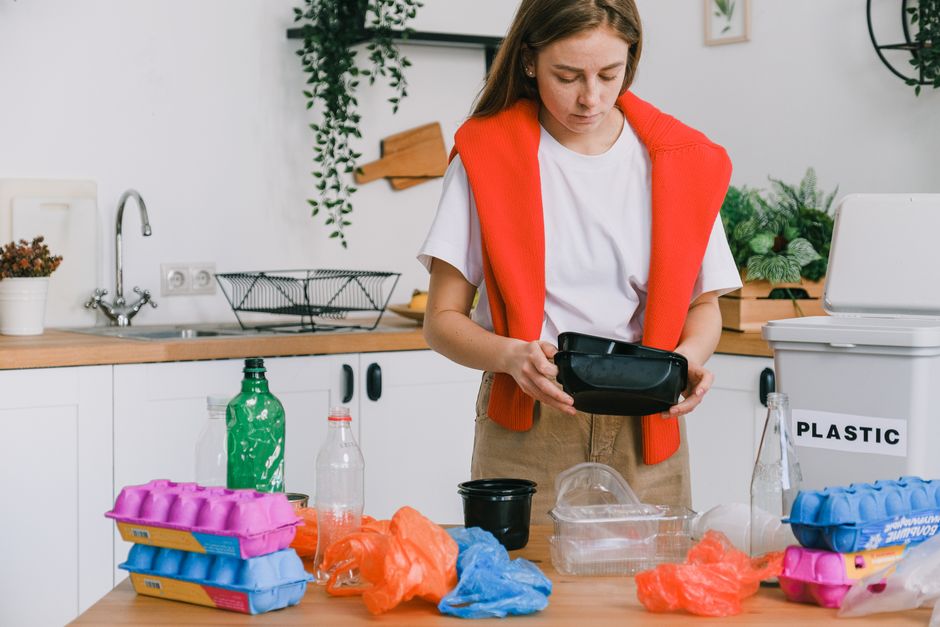 A young woman with shoulder-length brown hair, wearing a white t-shirt and a bright red jumper draped over her shoulders, stands in a modern, tidy kitchen with white cabinets and a wooden countertop. She is holding and inspecting a black plastic container, with her gaze focused on it. On the kitchen counter in front of her are various items related to waste sorting and disposal, including a clear plastic bottle, an empty black bowl, and several small colorful plastic bags in pink, orange, and blue. To her right, a white box labeled 'PLASTIC' is visible, partially out of the frame, indicating waste separation. There are also a green plastic soda bottle and a transparent bottle with a red cap among the clutter. The background features a stainless steel sink with a modern faucet, a wire dish rack, and decorative green indoor plants. The scene depicts an organized approach to managing recyclable waste, highlighting the importance of proper rubbish sorting and independent waste handling in a domestic setting, supporting services like those offered by Rubbish Clearance Lewisham for rubbish removal and waste duty of care compliance.