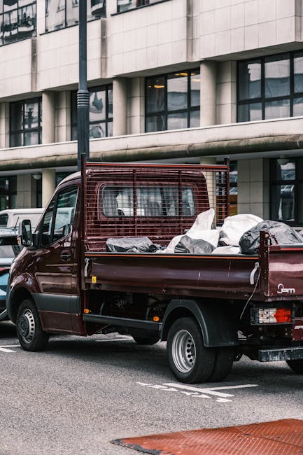 A small burgundy flatbed truck parked on a city street, loaded with various large black and grey rubbish bags and loose waste items. The truck features a metal cargo area with a protective mesh barrier at the front, separating the driver’s cabin from the load. It is positioned next to a modern building with large, rectangular windows and a light-colored facade. A tall black street lamp is visible beside the vehicle, casting minimal shadow, suggesting an overcast day with diffuse natural lighting. The scene depicts an environment where private waste collection or on-site clearance may be occurring, as the vehicle appears to be used for rubbish removal rather than regular municipal collection. The surface of the street is paved with asphalt, with white road markings indicating parking bays. The presence of other vehicles in the background further underlines an urban context, and the overall scene aligns with independent rubbish disposal activities often handled by private waste management services like Rubbish Clearance Lewisham, supporting the topic of alternative waste handling solutions outside standard council collections.